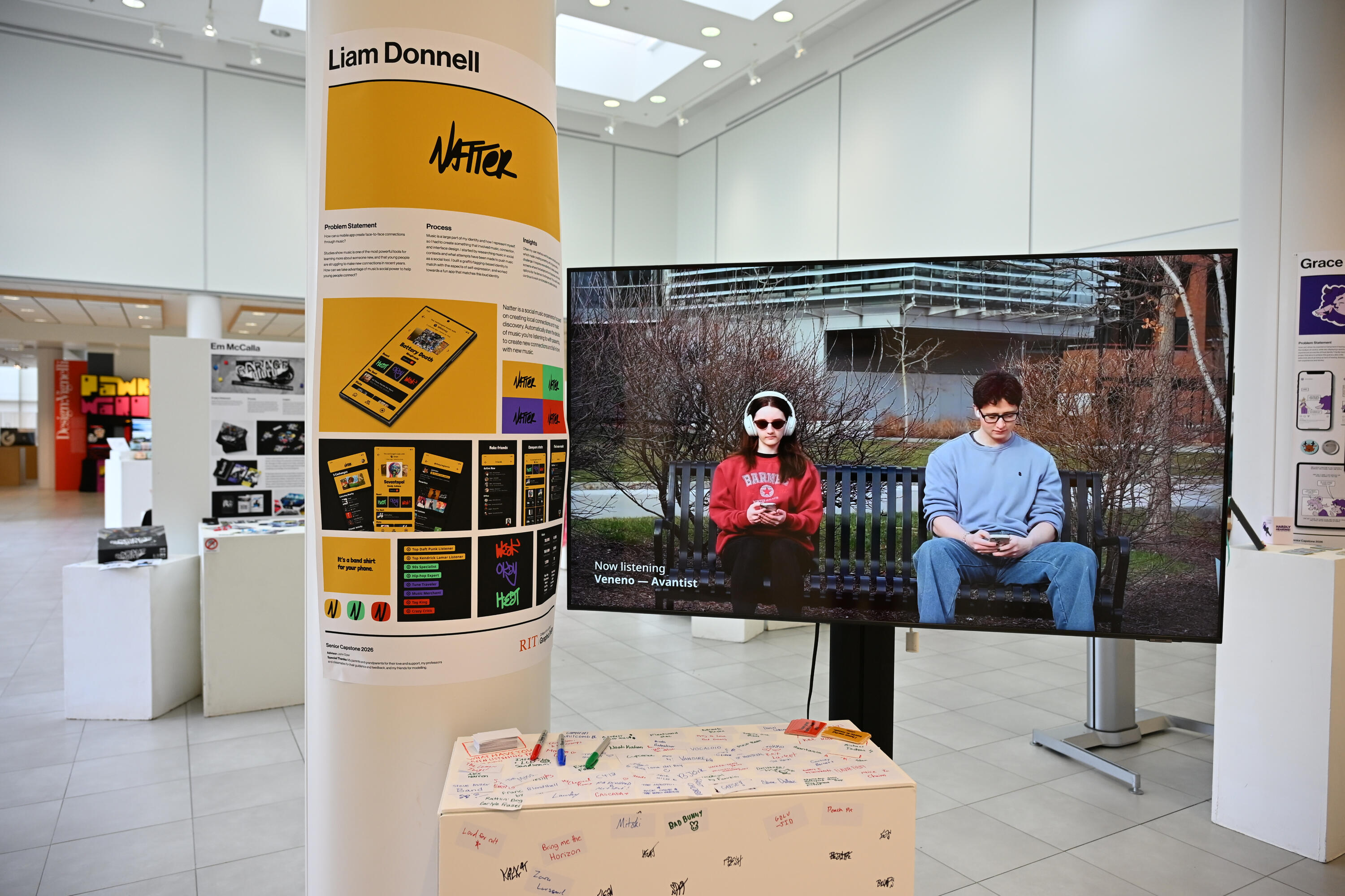 An exhibit in a gallery. To the left, a poster wrapped around a pillar, with various representations of the Natter app and identity. To the right, a large television with a video playing, where two students are sitting on a bench listening to music